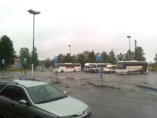 A cloudy day at a bus station, with several parked buses and a silver car in the foreground. Trees and streetlights are visible in the background.
