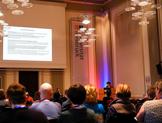 Die Aula der UNi Innsbruck ist gefüllt: Von hinten sieht man viele Menschen im Raum sitzen und eine KEG Sprecherin am Podium stehen.