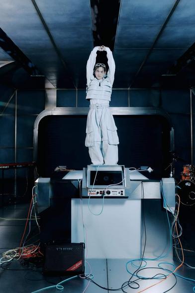 SUHO stands on top of a space tech-y center spaceship console with wires and monitors attached to it. He wears big bulky white cargo pants, long sleeve white shirt, bulky pocket-covered white vest, and white framed sunglasses on his head. He reaches both arms up above his head, making him look tall and long. He winks. A drumkit is obscured in a back corner.