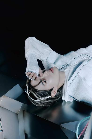 SUHO lays down on a metallic, angled platform and looks up to the camera with one hand blocking the light from one eye. He is wearing a bulky white outfit.