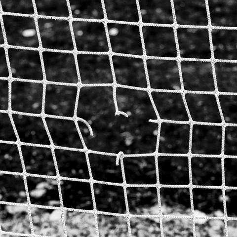 A close-up black and white photo of a torn soccer net with several strands loose against a dark, blurred field.