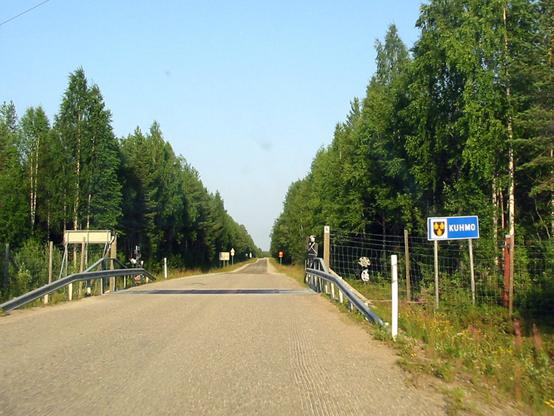 A rural road flanked by lush green trees under a clear blue sky. Municipality border sign for Kuhmo, Finland is on the side of the road. Behind it is a tall mesh fence, with some hubcaps hanging on it. At the border the road surface is interrupted by a section of metal grating intended to deter deer from crossing the border. A cutout silhouette figure of a human can be seen on the side of the road.