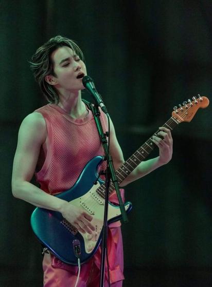 SUHO stands holding his blue and white electric guitar while singing into a microphone on a mic stand. He is wearing a pink satin jumpsuit with the top of it tied around his waist, showing a pink net tank top. His arms are bare. Half of his bangs are swooped over his face and the other half are swept back.
