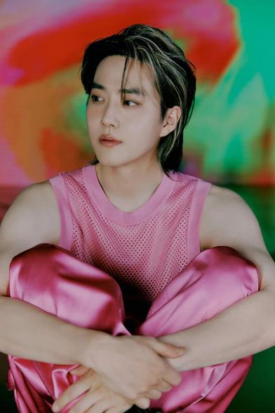 SUHO sits cross-legged on the ground, hugging his knees loosely. His pants are pink satin and his top is a pink net tanktop. His arms are bare and he looks off to the side. Behind him is a neon tie-dye colorful background.