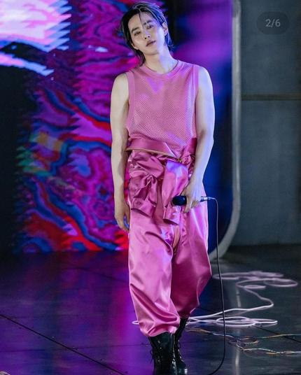 Full length view of SUHO in black stompy boots, a pink satin jumpsuit with the sleeves tied around his waist, and a pink tank top. He holds a microphone in one hand and walks toward the camera.