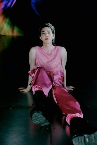 SUHO sits on the ground wearing his pink satin pants and pink tank top. He leans back on his hands and his legs are pointed at the camera with one bent slightly at the knee. He wears black stompy boots. His lips are parted with his tongue slightly sticking out.