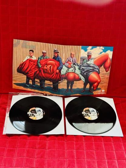 A double vinyl set of Amputechture by The Mars Volta is displayed on a red quilted surface. The album artwork in the background features a surreal painting of six men, each holding large, distorted human body parts wrapped in cloth, in a bizarre and dreamlike setting. The two black vinyl records are placed below the cover, each resting in a white paper sleeve. The center labels on the records also feature abstract artwork in a similar style to the cover.