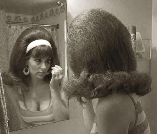 Eine Frau steht vor einem Badezimmerspiegel und trägt ein stark toupiertes, voluminöses Haar im Stil der 1960er Jahre. Sie trägt ein gestreiftes Oberteil, große runde Ohrringe und schminkt sich konzentriert die Augen. Ihr Haar ist nach hinten aufgebauscht und fällt in einer runden Welle nach außen. Die Szene wirkt retro und ist in Schwarz-Weiß gehalten, was den nostalgischen Eindruck verstärkt.
🖖