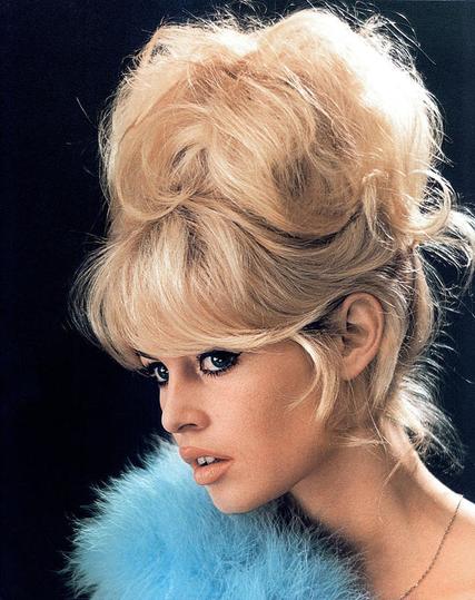 Porträt einer jungen Frau mit einer typischen 1960er-Jahre-Frisur: blond, voluminös und hochtoupiert, mit sanft geformten Strähnen, die sich nach oben und hinten legen. Sie trägt ein schlichtes Kleid und eine Perlenkette. Ihr freundliches Lächeln, das weiche Licht und der Hintergrund in pastelligem Grau-Grün erzeugen einen eleganten Vintage-Charakter.🖖