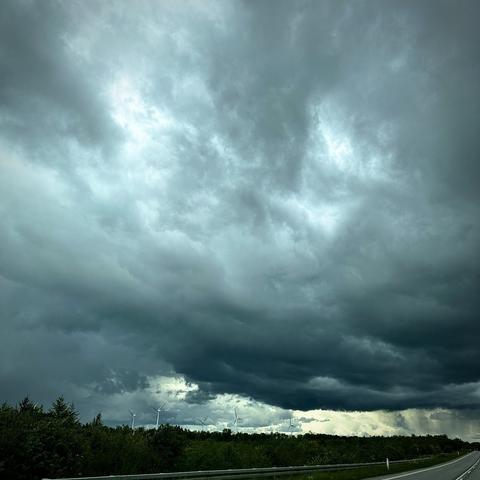 A dramatic sky featuring dark, cloudy weather with varying shades of gray. Wind turbines are visible in the background, surrounded by green trees and vegetation.
