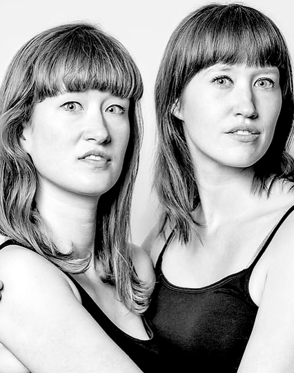 Two girls with straight, slightly longer hair with straight bangs. Both in black tank tops with thin straps.