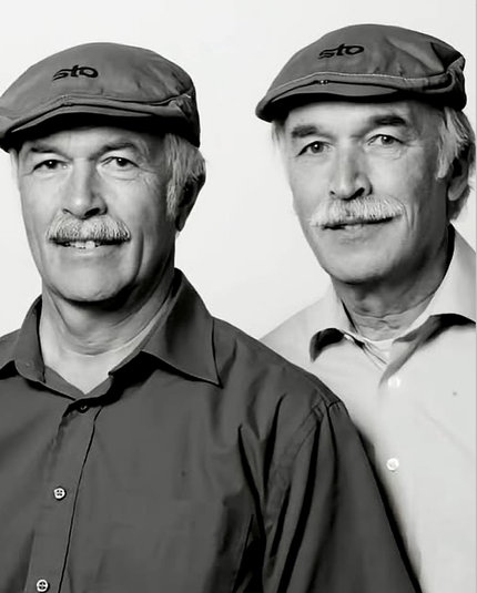 Two men in caps, one in a light shirt and the other in a dark one. Both with mustaches & gray hair.