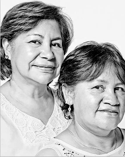 Two older women with short hair, they wear earrings & white blouses with lace.