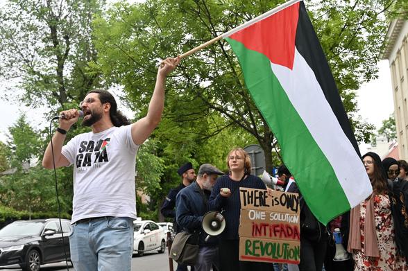 Osoba w koszulce z napisem "Save Gaza" z dużą flagą Palestyny wykrzykująca do mikrofonu
