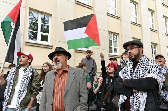 Grupa osób demonstrujących. W tle widoczne palestyńskie flagi. Dwie osoby ubrane w palestyńskie kefije.