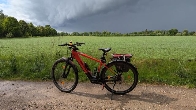Ein rotes Raleigh Dundee Mountainbike steht auf einem Schotterweg vor einem grünen Geld. Der Himmel zeigt eine dramatisch dunkle Wolkenwand während links im Bild noch ein kleines Stückchen blauer Himmel zu sehen ist.