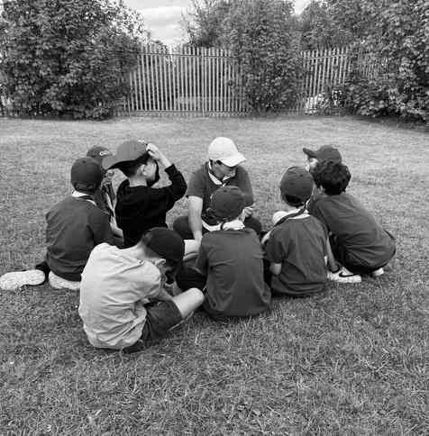 A black and white photo of a group of boys sitting on the lawn. There is a metal fence in the background. Most of the boys are wearing a cap.