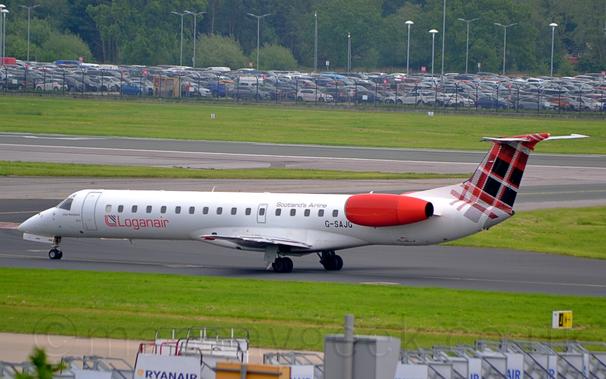 Side view of a white, twin engined jet airliner taxiing from right to left.
The plane has it's engines mounted on the sides of the rear fuselage, just below the tail.
It is mostly white, with red "LoganAir" titles on the lower rear fuselage, next to a red and black logo which resembles a red, upper-case letter "L" with a black triangle pointing to the upper right.
There is a red, black, and white tartan pattern on the tail.
Green grass fills most of the foreground, with a collection of mobile airstairs right at the bottom of the frame.
Taxiways and a runway lined with green grass, in the background lead up to a nearly full car park surrounded by a black metal mesh fence, with tall lighting poles scattered amongst the cars, with tall trees beyond.