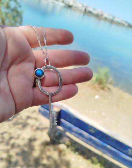 a silver key with a blue round gemstone. it is a pendant with black cord. it's holded on a hand with nature and sea background