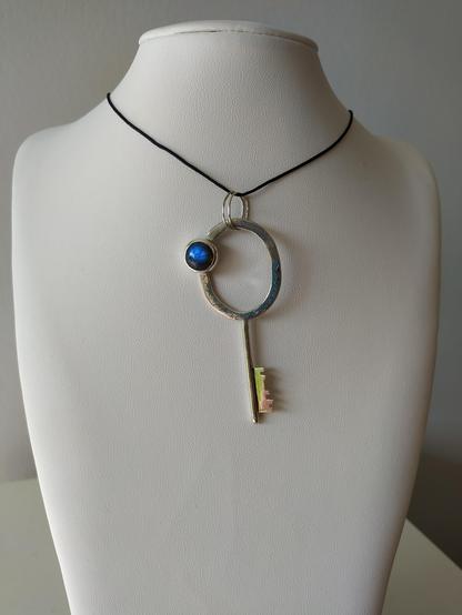 a silver key with a blue round gemstone. it is a pendant with black cord