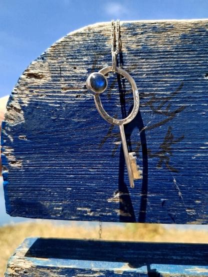 a silver key with a blue round gemstone. it is a pendant with black cord. it's hanged on a blue wooden piece