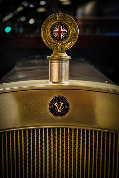 The image features a close-up view of the front of a vintage car, specifically focusing on the hood ornament and grille. The hood ornament is a circular emblem with a gold finish, prominently displaying the text "ROYAL AUTOMOBILE CLUB ASSOCIATE" and a central Union Jack flag. Below the ornament, the car's grille is visible, with vertical slats arranged in a diamond pattern. The car's brand is identified by a circular badge on the grille, which reads "VAUXHALL LUTON" with a stylized "V" and "L" in the center. The overall color scheme is a warm gold, with the emblem and badge providing a contrast against the darker background. The background is blurred, with hints of city lights, suggesting an urban setting.

Provided by @altbot, generated privately and locally using Ovis2-8B