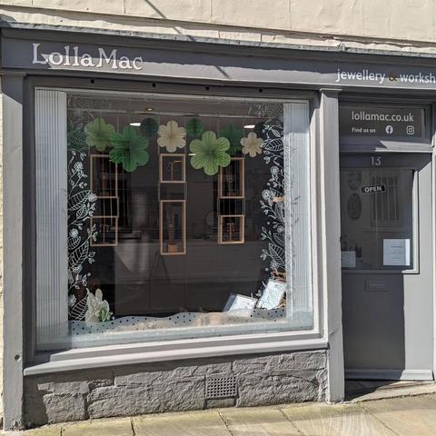 The front of the LollaMac shop. A large shop window in a stone building, with grey wooden frame, and the LollaMac logo above. The window display is hanging wooden boxes containing small jewellery items, with green paper decorations above, and hand-drawn leaves in white around the edge of the glass.