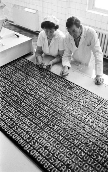 A black-and-white photograph showing two individuals, a woman in a hairnet and a man in a lab coat, inspecting a large metal plate covered with rows of embossed letters and symbols, likely related to printing or typography. They are actually cookies in letter form called “Russisch Brot”