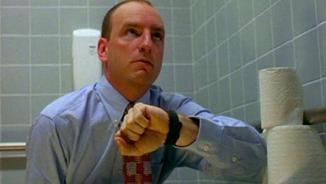 The image shows a person in a light blue dress shirt and a patterned tie, sitting in a bathroom with light gray tiled walls. The individual is looking intently at their wristwatch. To the right, there are several rolls of toilet paper stacked on a metal bar along the wall. The person appears focused, with a slightly furrowed brow and the setting suggests a public or office restroom.