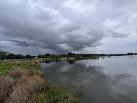 Photo of a stormy but clearing afternoon on Harris Creek and sky is filled with dark brooding cumulus clouds, with a bit of blue sky showing at the far horizon. Marsh is green with riotous late Spring growth. Creek water is rippled and reflects the sky and clouds.