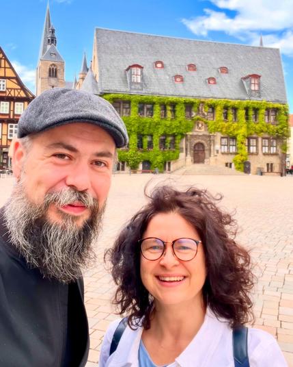 Ein lächelndes Paar posiert für ein Selfie vor dem Rathaus von Quedlinburg. Links ist ein Uhrenturm zu sehen. Der Himmel ist blau mit ein paar Wolken.