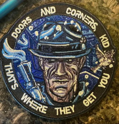 A round patch, face of Detective Miller and around the margin, the words, "Doors And Corners, kid. That's where they get you". A message from 'The Expanse'.