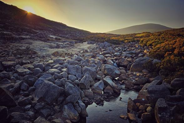 A wide shot of a rocky mountain valley at sunset. A small stream winds through the foreground, surrounded by large, dark grey boulders. The sun, a bright golden glow, peeks over the left mountain peak, casting a warm light across the upper right side of the image, illuminating some golden-orange gorse. The sky is a gradient of yellow, orange, and blue.