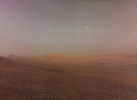 A grainy picture of Mars taken during astronomical twilight (the sun was 13° below the horizon). It shows a dimly lit reddish landscape with some hills at the horizon. Deimos, Mars' smaller moon shines with a small halo in the pale red-greyish sky above.