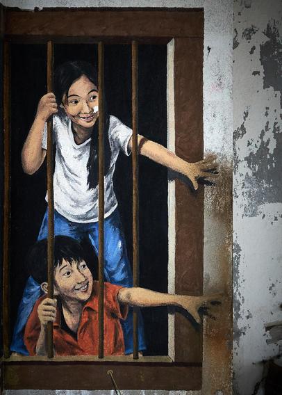 A mural showing two children, a boy and a girl, peering through wooden bars of a window. The girl, dressed in a white shirt, smiles as she holds the bars, while the boy, in a red shirt, leans forward, also smiling