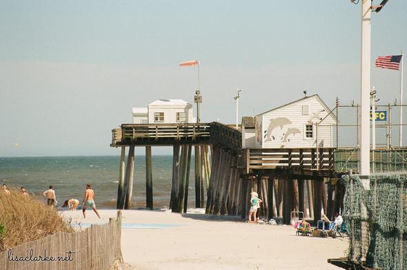 A group of people in bathing suits playing ball on the beach next to the Ocean City Fishing Pier. A few people, standing next to a pile of beach paraphernalia look on.