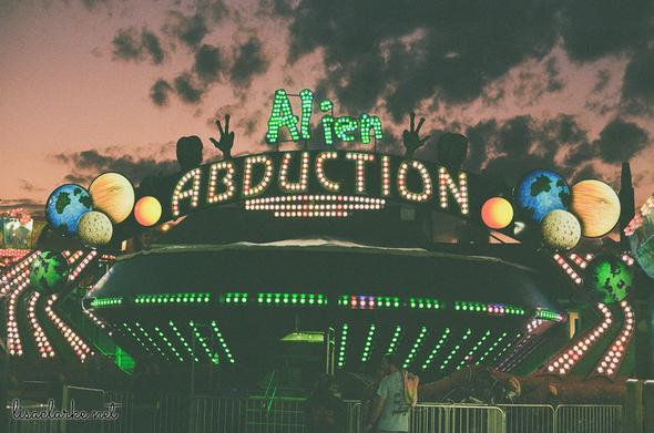Behind the lit-up "Alien Abduction" amusement, which is shaped like a UFO, is a dark, reddish-purple menacing sky with black clouds.