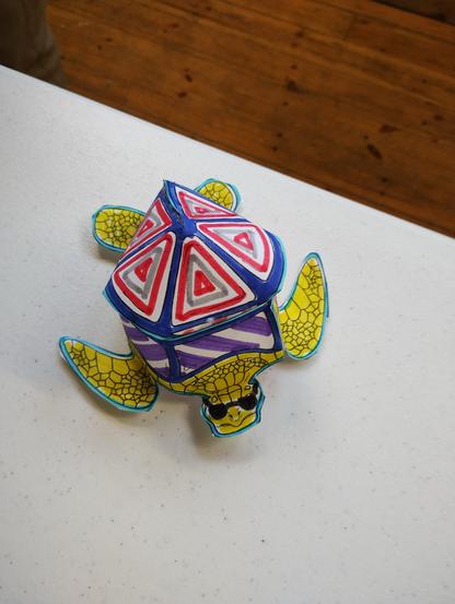 A 3 dimensional paper craft turtle, colourfully coloured in with felt pens with yellows reds blues and purples. It almost looks like a sweet you could eat.

It is on a white tabletop and a dark brown wooden floor can been seen behind.