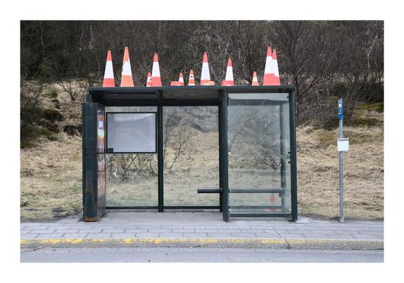 A bus stop shelter with glass walls and a bench inside. The roof of the shelter is lined with multiple bright orange and white traffic cones. The surrounding area consists of dry grass, sparse trees, and a small sidewalk.