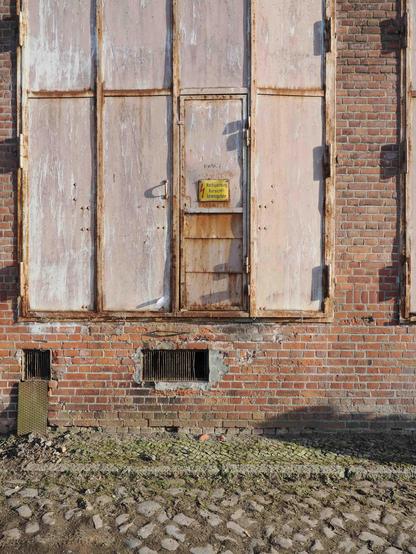 The image shows a brick wall with a large, weathered metal door. There is a smaller access door within it, marked with a yellow warning sign. The wall has two rectangular vents near the bottom, and the ground is a mix of cobblestones and dirt. The surfaces exhibit signs of rust, peeling paint, and general wear.