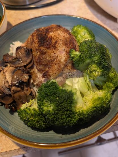 A bowl with rice covered in a chicken thigh, steamed broccoli and cooked mushrooms