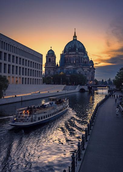 View on Berliner Dom and the Spree at sunset from a bridge, with the orange light from the sun reflecting on the river and making a nice contrast with the other tones in the image, which are more in the purple range. A touristic boat is advancing on the Spree making soft waves altering the reflection of the Dom.