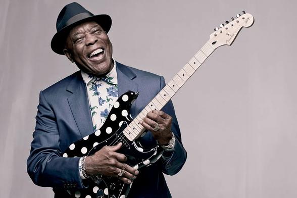 photograph of Buddy Guy, dressins a a suit and hat, holding his polka dot covered guitar