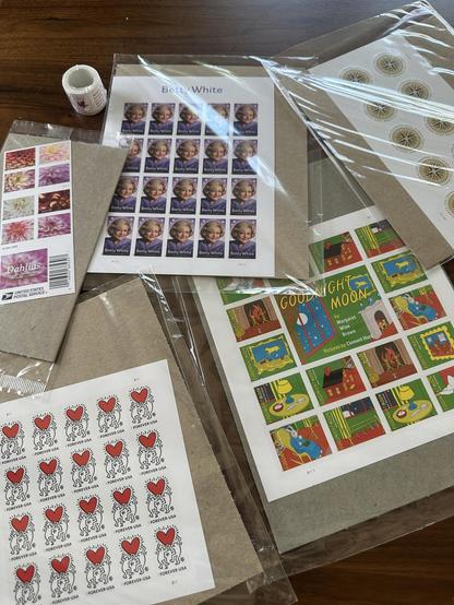 Photo of several sheets of US postage stamps laid out on a table. Designs include the Keith Herring love stamp, Betty White, dahlias, Goodnight Moon, a coil of 100 autumn leaves, postcard stamps and global stamps that feature a compass illustration.