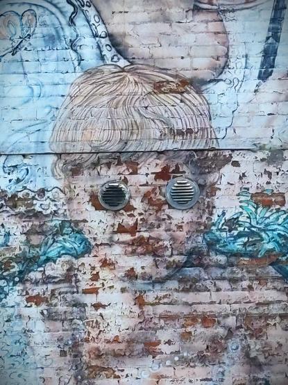 Photograph of a section of a wall mural that has seen better days. The paint is faded and peeling but you can still make out a portrait of a small child whose eyes have apparently been replaced with circular metal vents.