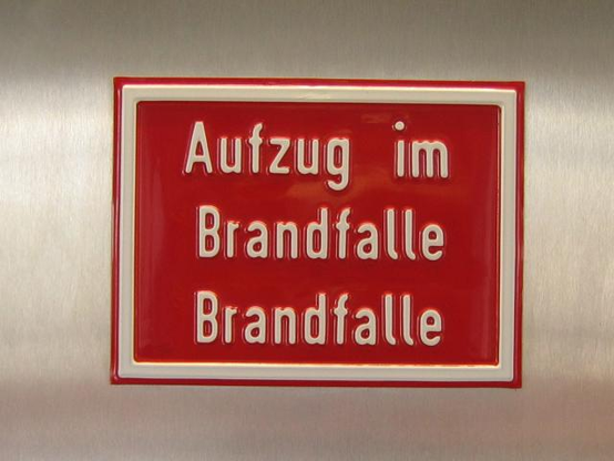 Ein rotes Aufzugschild vor metallenam Aufzugwandverkleidungsmeterials mit der Aufschrift:

Aufzug im 
Brandfalle
Brandfalle