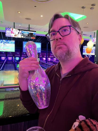 Ben Ramsey (the real one) holding a disco ball bowling pin in front of several bowling lanes. His tiger travel companion, Rawr, can be seen in the corner of the frame.