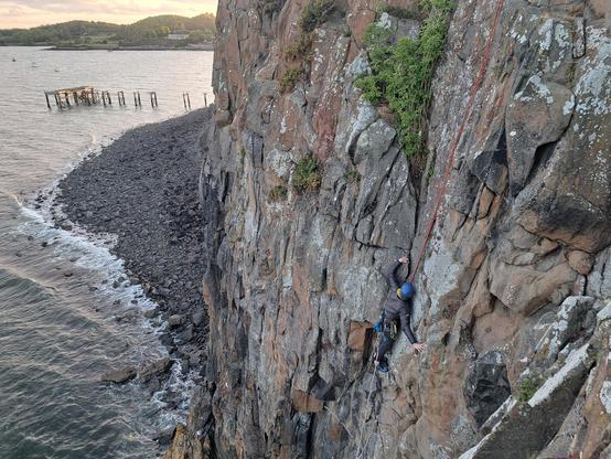 Image of climber spanning their arms wide as they move up an open groove. They are wearing a warm puffy jacket, with a blue helmet and are on a pink top rope. The rock is grey and orange-brown with many fractures. Below the sea washes onto a rocky beach. The distant sky is golden pink with the setting sun.
