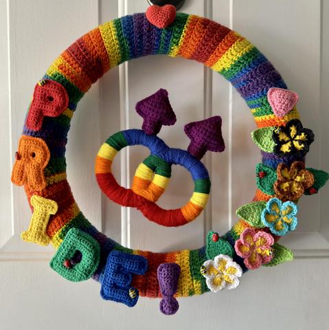 Male male pride wreath lighted with fairy lights. More than 20 individual pieces are assembled here to display all of the inclusive colors of the new inclusive pride flag! The Pride letters are done in the classic Pride colors and then the edge of the flowers shows the additional colors! A small pink Pride triangle is also there to denote remembrance of those lost in the holocaust. There are multiple wooden ladybugs and bees just to give the whole wreath a little life! 13” inches round and 100% handmade!