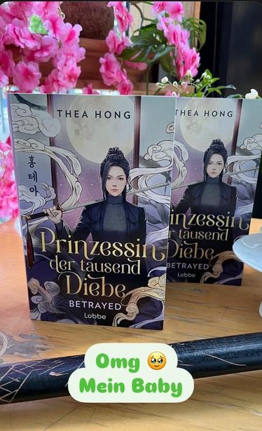 The image features two copies of the book "Prinzessin der tausend Diebe" by Thea Hong, displayed on a wooden surface surrounded by pink flowers. A decorative black object is also visible, accompanied by a caption that expresses excitement.